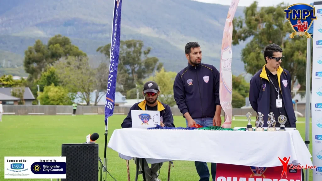 Crew at a ground-side desk with speakers and trophies, hills behind